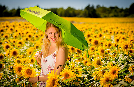 Eine Frau steht in mitten eines Sonnenblumenfeldes und trägt dabei einen Regenschirm.